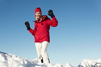 Femme debout dans la neige, vêtue d'une parka vert foncé, capuche en fourrure, tenant une tasse de café et un smartphone.