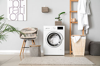 Utilisez correctement les accessoires pour entretien du linge : dans une salle de bain, il y a un lave-linge remplie, à gauche une chaise avec des coussins décoratifs et à droite une haute étagère en bois ouverte avec un panier à linge devant.