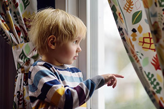 Un petit garçon regarde avec intérêt par une fenêtre à laquelle sont accrochés des voilages colorés.