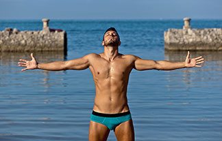 Sur une plage, un jeune homme sportif portant un slip pour hommes aux bras tendus apprécie le soleil.