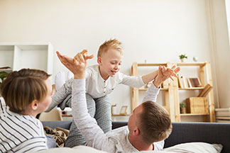 Une jeune famille est allongée sur le lit, ils sont habillés avec des survêtements d’intérieur et le père fait voler son petit garçon sur ses jambes « comme un avion ».