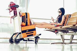 Set de valises & accessoires de voyage pour femmes : une jeune femme est assise à l'aéroport, en train de regarder son smartphone. Ses jambes sont allongées sur son set de valises, lui-même posé dans un chariot à bagages.