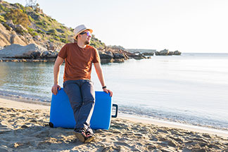 Sur une plage, un jeune homme portant un chapeau est assis une valise bleue pour hommes.