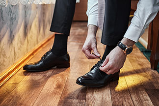 Gros plan sur les jambes d'un homme portant un pantalon de tailleur noir, des chaussures en cuir noires et des chaussettes pour hommes noires, il est assis sur son lit et en train de lacer ses chaussures.