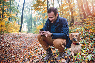 Un homme portant un manteau mi-saison et une paire de bottes pour hommes imperméables est agenouillé dans une forêt aux couleurs d'automne et utilise son smartphone, son chien est couché à côté de lui.