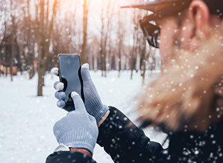 Casquettes et gants pour hommes à détails : sur un fond de paysage hivernal enneigé, un homme est debout, en train d’utiliser son smartphone avec ses gants avec ses gants aux mains.