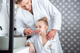 Dans une salle de bains, un père et sa fille, portant chacun un peignoir blanc, le père est en train de brosser les dents de sa fille.