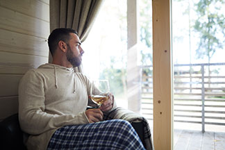 Un jeune homme portant un survêtement d'intérieur pour hommes composé d'un pantalon à carreaux et d'un pull à capuche de couleur claire est assis à la fenêtre et est en train de boire un verre de vin.