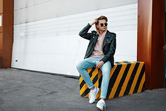 Chaussures basses pour hommes pour l'été : un jeune homme portant un jean, un sweat à capuche et des baskets blanches est assis, en train de se détendre sur un cube de béton rayé noir et jaune devant un garage.