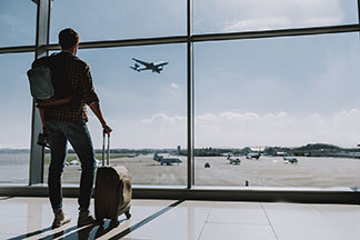 Un jeune homme positionné de dos, se tient près de la baie vitrée d'un aéroport. Il tient fermement une petite valise pour hommes qui se trouve à côté de lui.