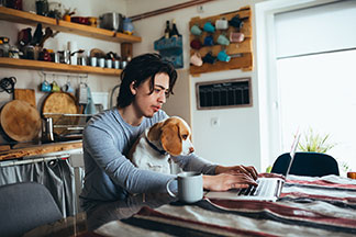 Un jeune homme portant un survêtement d'intérieur pour hommes travaille sur son ordinateur portable à la table de sa cuisine, avec son chien sur les genoux.