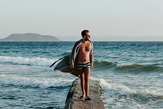 Un jeune homme portant un maillot de bain, des lunettes de soleil et un peignoir pour hommes de couleur beige ouvert est en train de marcher sur un ponton de pierre allant vers la mer.