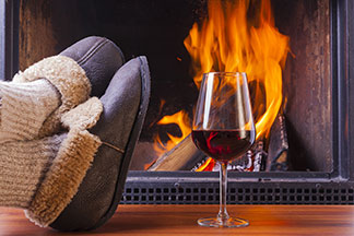 Chaussons pour hommes pour l'hiver : gros plan sur des chaussons confortables devant une cheminée. Un verre de vin rouge est posé à côté.