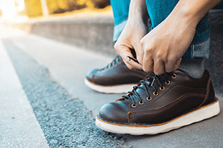 Gros plan sur des chaussures basses pour hommes en coloris marron, avec un homme assis sur une marche d'escalier en train de lacer ses chaussures.