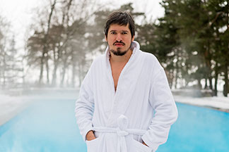 Un homme barbu vêtu d'un peignoir pour hommes blanc se tient à l’extérieur, devant une piscine fumante, les mains dans les poches.