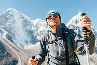 Un homme porte une veste de ski rouge, des accessoires de sport pour hommes comme des gants et bâtons de ski, des lunettes de ski et se tient sur une piste dans un paysage de montagne enneigé.