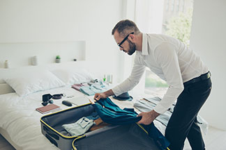 Un homme portant une chemise blanche et un pantalon noir, prépare sa valise et accessoires de voyage, la valise est ouverte sur le lit.