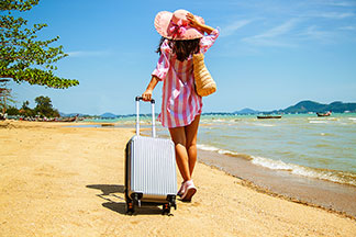 Valises & accessoires de voyage de qualité pour femmes : une jeune femme portant un chapeau tire son trolley en métal sur une plage de sable fin.