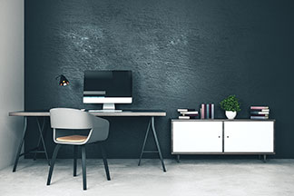 Un bureau avec surface de travail et pieds en métal foncé disposé contre un mur sombre - à côté, une commode blanche sur laquelle sont posés des livres et une plante verte.