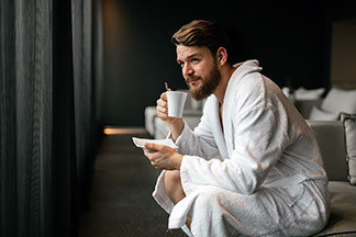 Assis sur le bord du lit, un jeune homme en peignoir pour hommes blanc déguste un thé dans une tasse blanche.