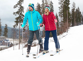 Les bons vêtements de ski pour les pistes : un jeune couple, chacun portant une tenue de ski en deux pièces, se tient sur une piste de ski, souriant avec joie.