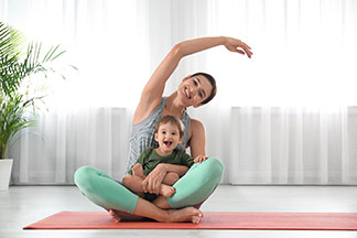 Vêtements de yoga de qualité supérieure : une jeune mère est assise sur son tapis de yoga et lève le bras au-dessus de sa tête, son fils est assis sur ses genoux.