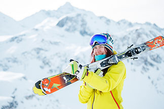 Vêtements de ski en softshell : une jeune femme joyeuse en veste softshell jaune porte ses skis sur l'épaule avec un paysage de haute montagne en arrière-plan.