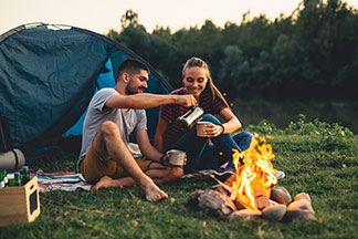 Le bon équipement : un jeune couple en tenue de plein air est assis de bonne humeur devant leur tente au coin du feu et boit du café.