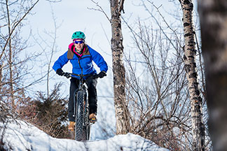 Une jeune femme fait une montée en vélo dans un paysage hivernal, vêtue d'une veste bleue et d'un pantalon noir.