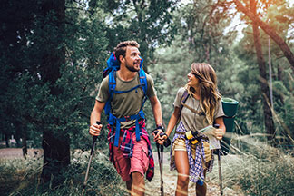 Un homme et une femme, portant tous deux des vêtements d'extérieur, marchent côte à côte. Tous deux portent des shorts, des t-shirts, des bâtons de randonnée ainsi qu’un sac à dos.