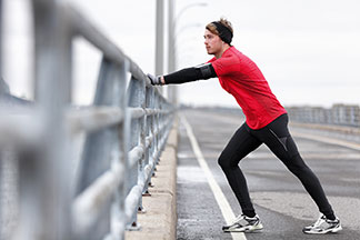 Un jeune homme sportif en sous-vêtement fonctionnel à manches longues, sur lequel il porte une chemise rouge, fait des exercices d'étirement sur un pont pendant sa séance d'entraînement à la course.