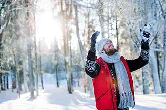 Un homme barbu, portant une casquette, des gants et une écharpe d'hiver, se tient dans une forêt enneigée et tend les bras vers le ciel.