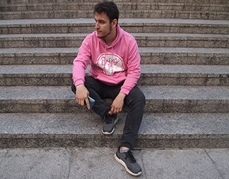 Un jeune homme tenant un smartphone est assis sur un escalier de pierre, vêtu d'un sweat-shirt rose à capuche.