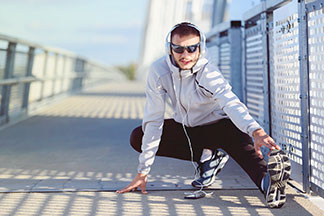 Des vestes de sport hommes pour toutes les situations : un jeune homme en veste de course blanche fait des exercices d'étirement sur un pont avec des écouteurs.