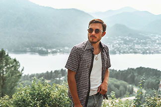 Un jeune homme vêtu d'une chemise manches courtes à carreaux, portée ouverte, se tient devant un lac avec des montagnes en arrière-plan.