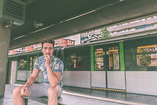 Un jeune homme en chemise manches courtes avec un motif floral et des pantalons courts est assis sur un banc dans un arrêt de train de banlieue (RER).