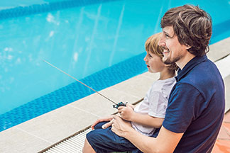 Un père portant un polo bleu est assis avec son fils au bord d'une piscine. Tous deux manœuvrent ensemble un bateau télécommandé.