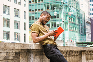Un jeune homme portant un jean avec une ceinture et une chemise beige manches courtes est debout en train de lire, appuyé contre un pont de pierre.