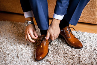 Des chaussures hommes pour le bureau : gros plan des pieds d'un homme en costume bleu, assis sur son lit et nouant les lacets de ses chaussures en cuir marron.
