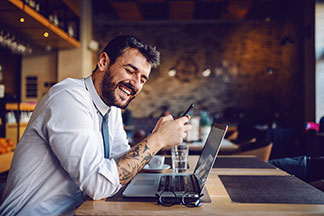 Un homme barbu et tatoué est assis devant son ordinateur portable dans un café, vêtu d'une chemise hommes bleu clair, et il rit.