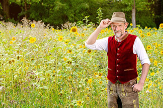 Gilets traditionnels hommes : un homme portant un pantalon en cuir et un gilet rouge se tient devant un champ de tournesols, serrant son chapeau.