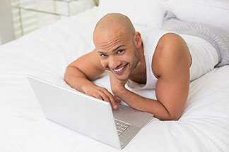 Un homme portant un sous-vêtement blanc et un long pantalon de pyjama est allongé dans son lit et travaille sur son ordinateur portable.