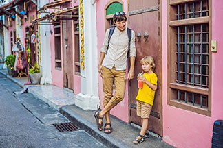Des shorts hommes dans toutes les variations : un père en short 3/4 se tient debout, appuyé contre un mur de la maison, avec son fils portant un short à longueur de genou.