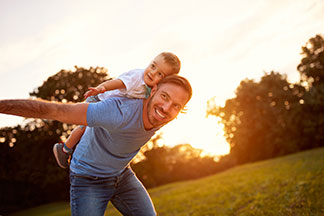 Pantalons hommes en qualité Tchibo : un père souriant en jeans et avec un t-shirt bleu balance son jeune fils dans un parc, avec les bras bien étendus sur son dos.