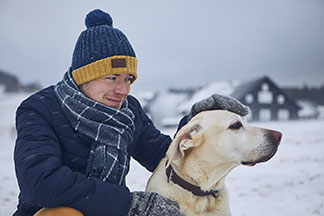L'accessoire idéal pour l'hiver : un jeune homme portant une écharpe, une casquette et des gants s'accroupit pour caresser son labrador blanc dans un paysage hivernal enneigé.