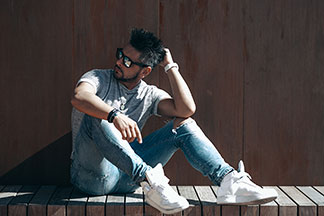 Chaussures hommes pour les loisirs : Un jeune homme portant des baskets blanches, un jean, une chemise grise et des lunettes de soleil est assis devant un mur de couleur marron.