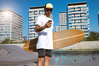 Un skater portant des lunettes de soleil et une casquette se tient dans un skate park avec une chemise surdimensionnée et son longboard à la main, en regardant son téléphone portable.