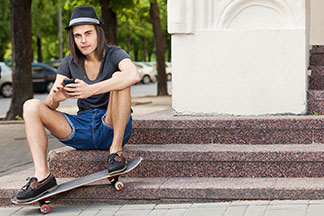 Un patineur élégamment vêtu, portant un chapeau sombre et un short en denim, est assis de manière décontractée sur un escalier de marbre, utilisant son smartphone.