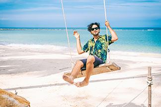 Look décontracté : un homme barbu en chemise hawaïenne manches courtes est assis sur une balançoire sur la plage. En arrière-plan, on peut voir la mer.