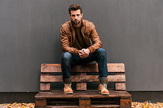 La meilleure qualité pour vos chaussures hommes : un homme portant des bottes marron et une veste en cuir marron est assis à l'extérieur sur un banc fait de palettes en bois.